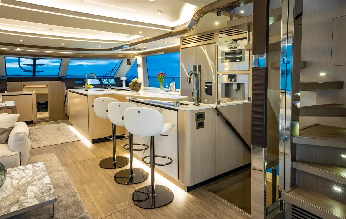 Luxurious yacht interior featuring a modern kitchen, three white bar stools, marble coffee table, and large windows with ocean views.